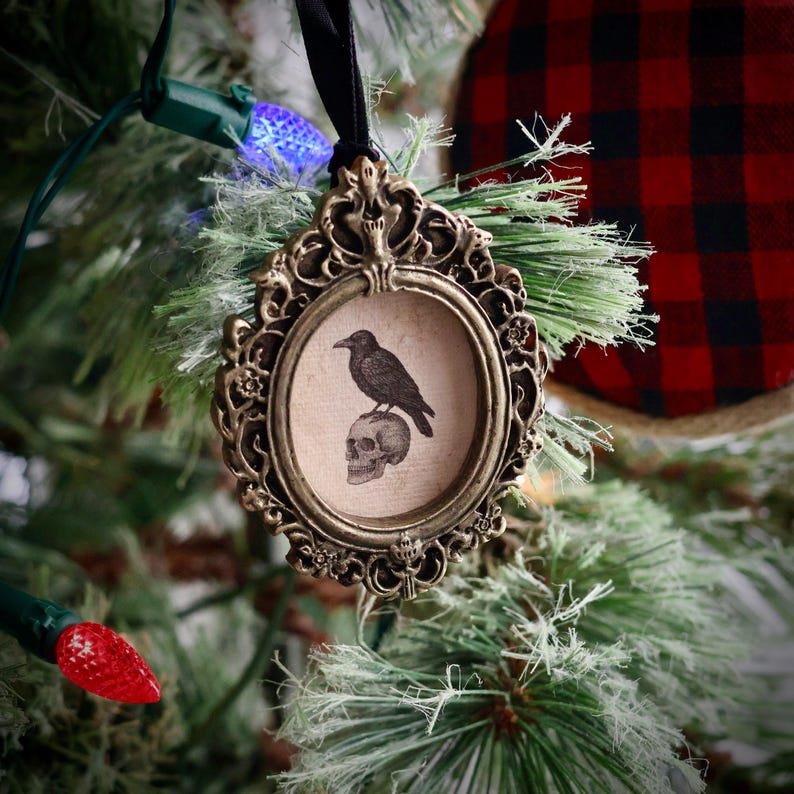 May include: Ornate, antique-style Christmas ornament with a black raven perched atop a skull inside an oval frame. The frame is bronze-colored and has intricate detailing. A black ribbon is attached for hanging. The ornament is displayed on a Christmas tree.