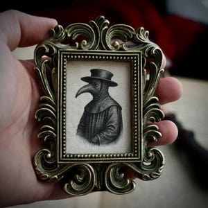 Puede incluir: Un pequeño marco de latón ornamentado de estilo antiguo contiene una ilustración en blanco y negro de un médico de la peste. El médico lleva un sombrero y una máscara con pico largo. El marco tiene intrincados adornos.