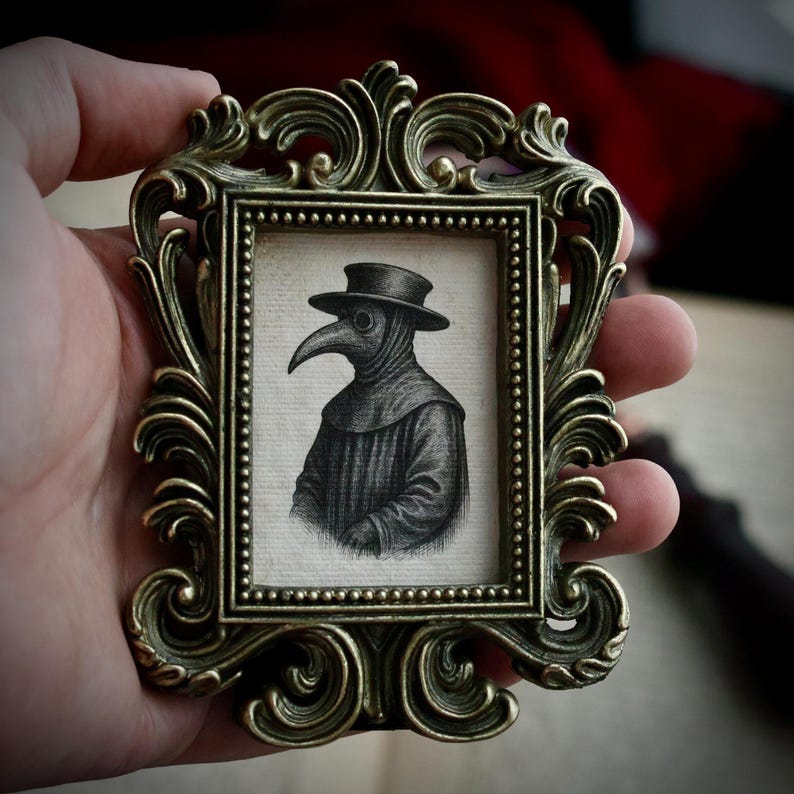 May include: A small, ornate, antique gold-colored frame holds a black and white illustration of a plague doctor. The doctor is wearing a hat and a long-beaked mask. The frame has detailed scrollwork.