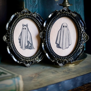 May include: Two oval-shaped black ornate frames containing illustrations of ghosts on a light beige background. The left frame features a ghost wearing a small bow on its head, while the right frame displays a ghost without any accessories.