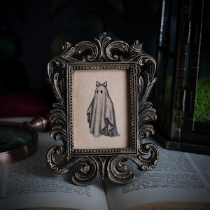Peut inclure: Dessin au crayon d'un fant&ocirc;me avec un n&oelig;ud papillon, encadr&eacute; dans un cadre orn&eacute; de couleur bronze. Le cadre est pos&eacute; sur un livre ouvert. Le fant&ocirc;me a deux yeux noirs et est drap&eacute; dans un drap. Le cadre est d&eacute;coratif.