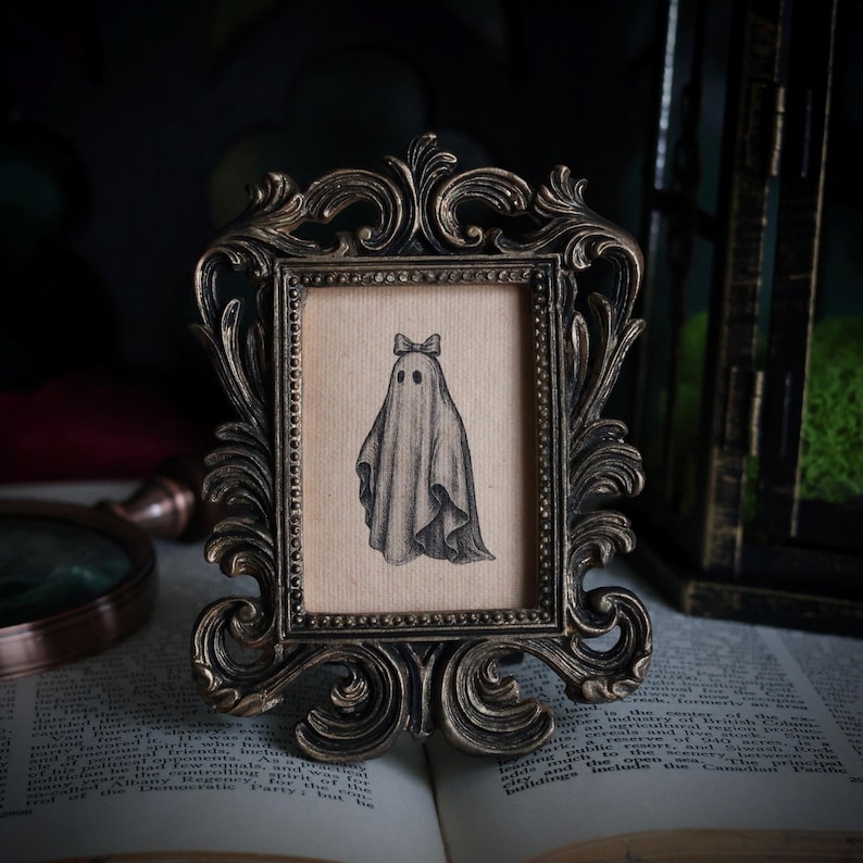 Peut inclure: Dessin au crayon d'un fant&ocirc;me avec un n&oelig;ud papillon, encadr&eacute; dans un cadre orn&eacute; de couleur bronze. Le cadre est pos&eacute; sur un livre ouvert. Le fant&ocirc;me a deux yeux noirs et est drap&eacute; dans un drap. Le cadre est d&eacute;coratif.