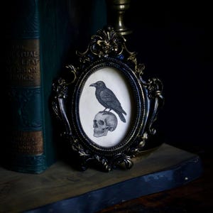May include: An oval-shaped black and gold ornate frame containing a cream-colored print of a black raven perched atop a human skull. The frame features intricate floral and scrollwork detailing along its edges, surrounding the central illustration.