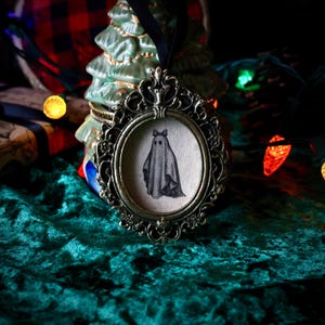 May include: An oval, antique-style Christmas ornament with a black and white ghost illustration. The ghost wears a bow. The ornament is framed in a bronze-colored metal with an ornate design and hangs from a black velvet ribbon.