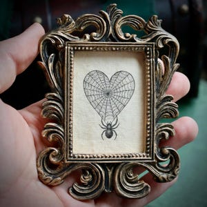 May include: Ornate, antique-style picture frame in gold with a heart-shaped spiderweb design and a spider illustration. The frame has intricate scrollwork and a vintage aesthetic.