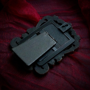 May include: A dark, ornate picture frame with a textured back and a hinged stand. The frame is dark brown with decorative carvings. The back is a dark gray color. The frame is resting on a deep red fabric.