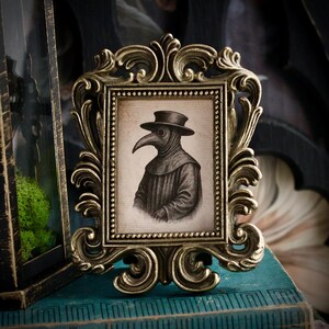 May include: An ornate, antique-style picture frame in gold tones. The frame holds a black and white illustration of a plague doctor wearing a hat and beak-like mask. The frame is placed on a teal-colored surface.