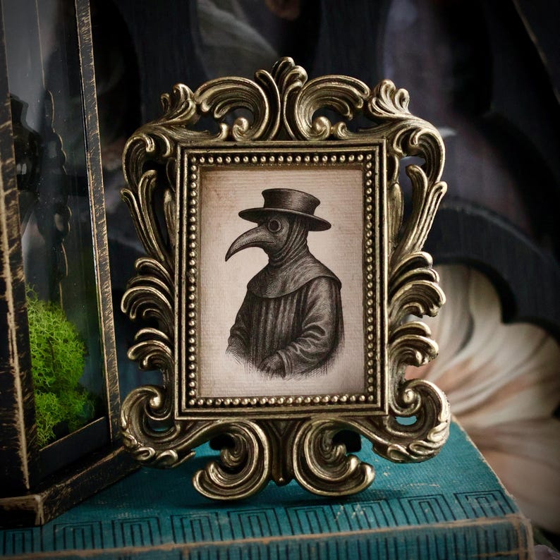 May include: An ornate, antique-style picture frame in gold tones. The frame holds a black and white illustration of a plague doctor wearing a hat and beak-like mask. The frame is placed on a teal-colored surface.