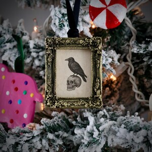 May include: A framed ornament with a black raven perched on a skull. The frame is ornate with a gold finish, and the background is a snowy Christmas tree. A black ribbon is attached for hanging.
