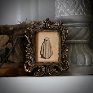 Peut inclure: Une petite impression encadr&eacute;e d'un fant&ocirc;me. L'&oelig;uvre d'art est dans un cadre dor&eacute; orn&eacute;. Le fant&ocirc;me est repr&eacute;sent&eacute; comme un drap&eacute; avec deux trous noirs pour les yeux. Le cadre a un design baroque antique.