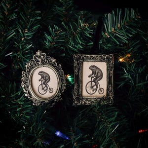May include: Two antique-style Christmas ornaments. One is oval, the other rectangular, both with ornate frames. Each features a sepia-toned image of a frog riding a penny-farthing bicycle. The ornaments are set against a backdrop of green Christmas tree branches.