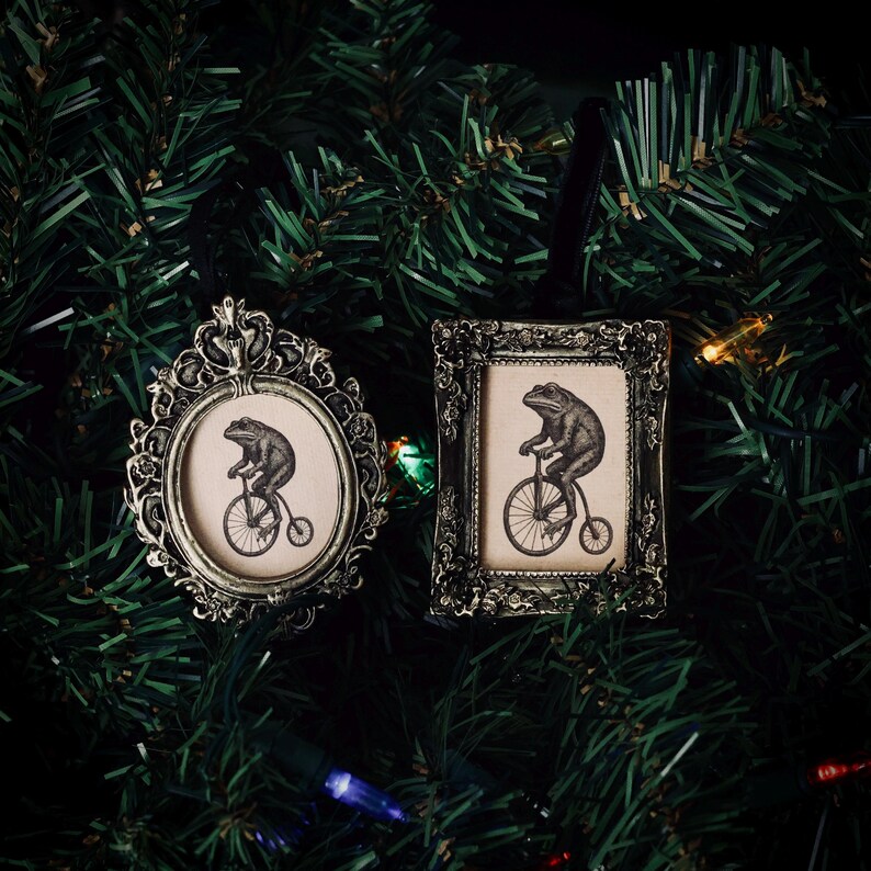 May include: Two antique-style Christmas ornaments. One is oval, the other rectangular, both with ornate frames. Each features a sepia-toned image of a frog riding a penny-farthing bicycle. The ornaments are set against a backdrop of green Christmas tree branches.