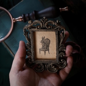 May include: A small, ornate, antique-style picture frame in a bronze finish. The frame holds a sepia-toned print of a rabbit seated on a chair. The frame is held in a person's hand, with a magnifying glass and other objects in the background.