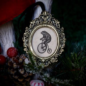 May include: An ornate, gold-colored framed ornament with a black and white illustration of a frog riding a penny-farthing bicycle. The oval-shaped image is set against a cream-colored background. A black ribbon is attached for hanging. The ornament is surrounded by festive greenery.