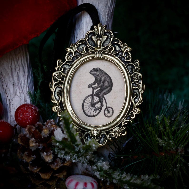 May include: An ornate, gold-colored framed ornament with a black and white illustration of a frog riding a penny-farthing bicycle. The oval-shaped image is set against a cream-colored background. A black ribbon is attached for hanging. The ornament is surrounded by festive greenery.