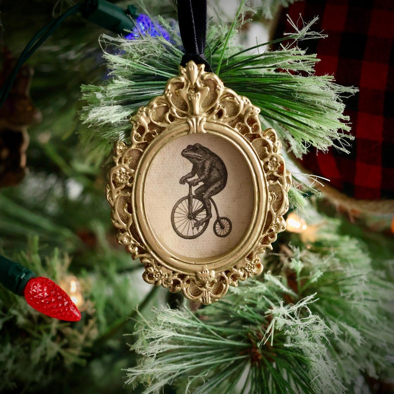 May include: A gold-colored, ornate Christmas tree ornament. The ornament features a circular frame with a black and white illustration of a frog riding a bicycle. The ornament is hanging from a black ribbon and is surrounded by green pine needles.