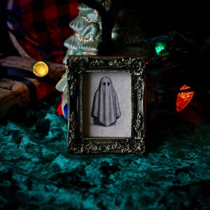 May include: A framed artwork featuring a gray ghost illustration. The artwork is in a decorative, ornate, dark-toned frame. The background includes a Christmas tree ornament and a green velvet surface.