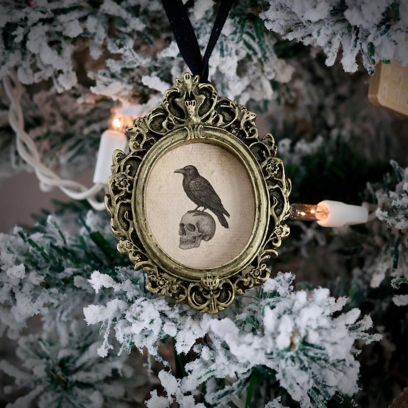 May include: Ornate, antique-style Christmas ornament with a black raven perched on a skull inside an oval frame. The frame is gold-toned with intricate detailing, suspended by a black ribbon. The background shows a snow-covered Christmas tree.