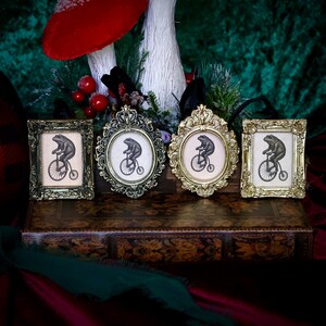 May include: Four ornate picture frames with a whimsical frog riding a bicycle illustration. The frames are in various shapes and metallic finishes, including gold and bronze. The artwork is set against a neutral background.