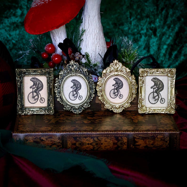 May include: Four ornate picture frames with a whimsical frog riding a bicycle illustration. The frames are in various shapes and metallic finishes, including gold and bronze. The artwork is set against a neutral background.