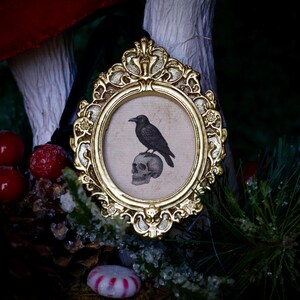 May include: A framed print featuring a black raven perched atop a skull. The print is set within an ornate, gold-colored oval frame. The background includes red berries, pine needles, and a candy cane.