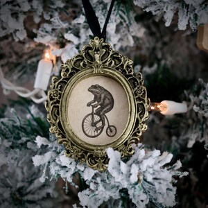 May include: Ornate gold-colored Christmas ornament with a black ribbon. The ornament features a circular image of a frog riding a penny-farthing bicycle. The ornament is hanging on a snow-covered Christmas tree branch.