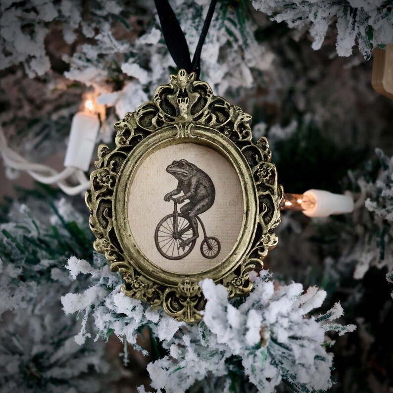 May include: Ornate gold-colored Christmas ornament with a black ribbon. The ornament features a circular image of a frog riding a penny-farthing bicycle. The ornament is hanging on a snow-covered Christmas tree branch.