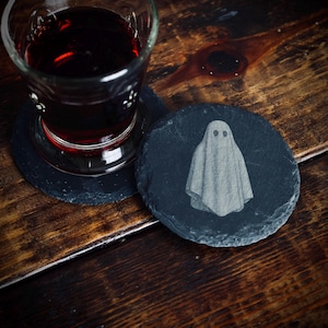 May include: A glass of red liquid rests on a black coaster, beside a round black slate coaster featuring a ghost design. The coasters are on a dark wooden surface. The ghost design has two eye holes and a draped sheet.