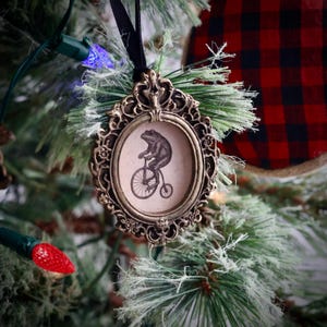 May include: Ornate, antique-style Christmas tree ornament. The bronze-colored frame surrounds a circular image of a frog riding a penny-farthing bicycle. The ornament hangs from a black ribbon and is set against a backdrop of a decorated Christmas tree.