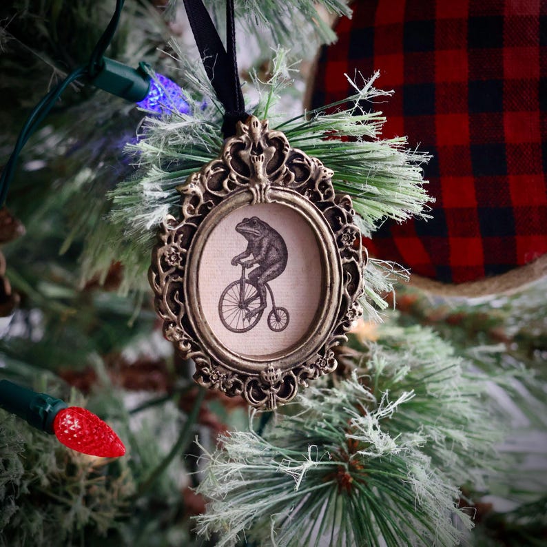May include: Ornate, antique-style Christmas tree ornament. The bronze-colored frame surrounds a circular image of a frog riding a penny-farthing bicycle. The ornament hangs from a black ribbon and is set against a backdrop of a decorated Christmas tree.