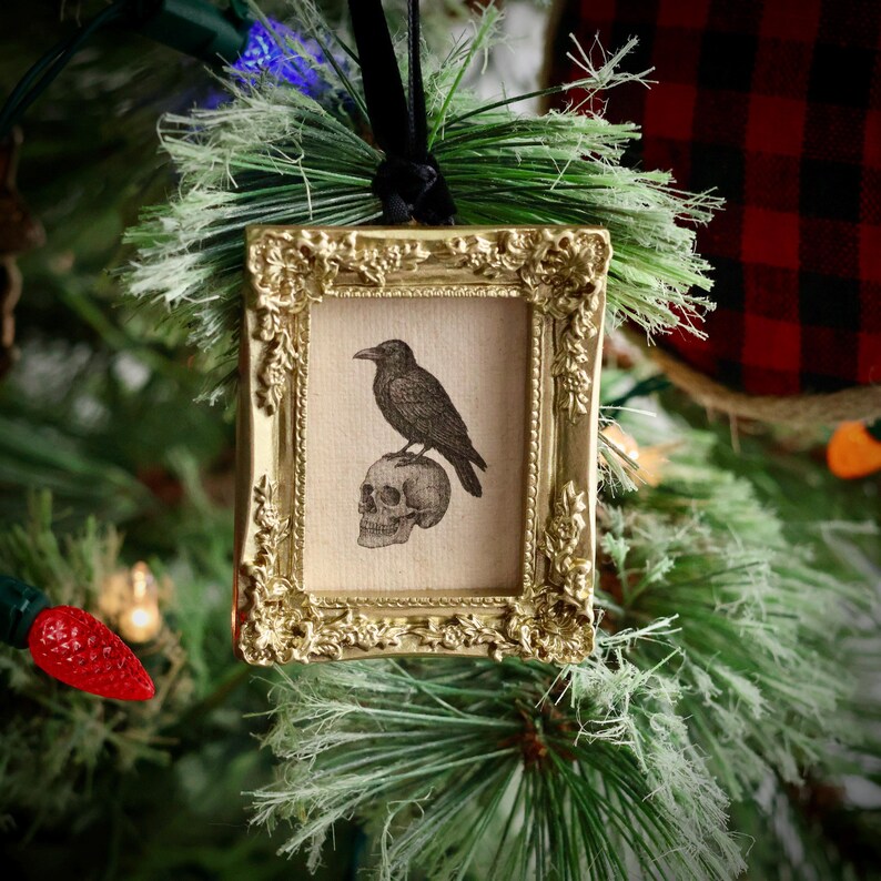 May include: A gold-colored framed ornament with a black raven perched on a skull. The ornament is hanging on a Christmas tree with green needles and red and blue lights. The frame has an ornate, vintage design.