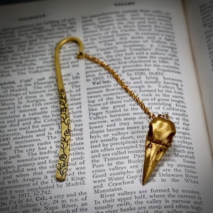 May include: A gold-colored metal bookmark with a raven skull pendant. The bookmark has a floral design and a chain connecting it to the skull. The bookmark is resting on an open book.