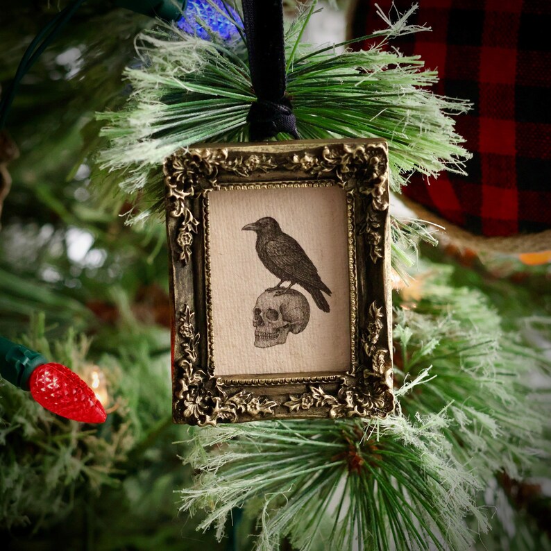 May include: Ornate, antique-style Christmas ornament featuring a framed image of a raven perched atop a skull. The frame is gold-toned with intricate detailing, and the image has a vintage aesthetic. The ornament hangs from a black ribbon.