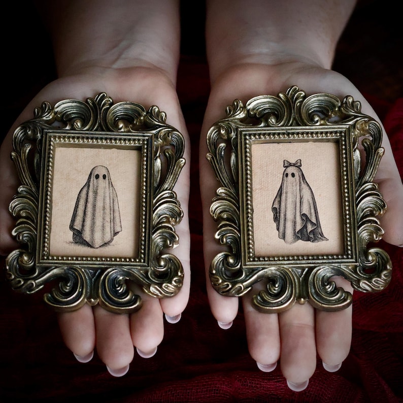 Peut inclure: Deux petits cadres dor&eacute;s antiques orn&eacute;s, chacun contenant un dessin s&eacute;pia d'un fant&ocirc;me. Un fant&ocirc;me porte un n&oelig;ud. Les cadres sont tenus dans des mains ouvertes sur un fond rouge fonc&eacute;.