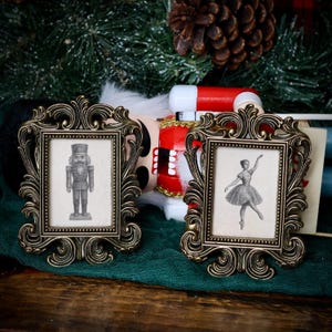 May include: Two ornate, antique-style picture frames with detailed gold-colored borders. Each frame holds a black and white image: one of a nutcracker soldier and the other of a ballerina. The frames sit on a green cloth.
