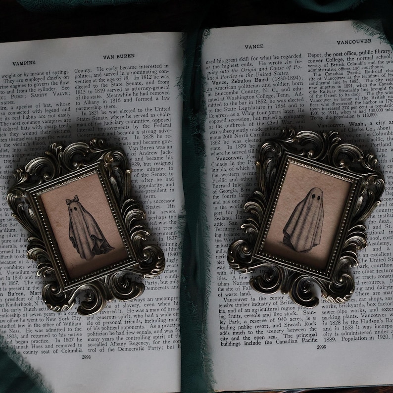 Peut inclure: Deux images encadr&eacute;es de style vintage avec une illustration de fant&ocirc;me. Le fant&ocirc;me est blanc avec un visage simple, portant un n&oelig;ud. Les cadres ont des d&eacute;tails en m&eacute;tal fonc&eacute; et orn&eacute;. Le fond est un livre ouvert.