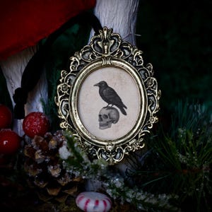 May include: Ornate gold-colored framed ornament featuring a black raven perched atop a skull. The oval image has a vintage aesthetic. A black ribbon is attached for hanging. The background includes red berries, pine needles, and a pine cone.