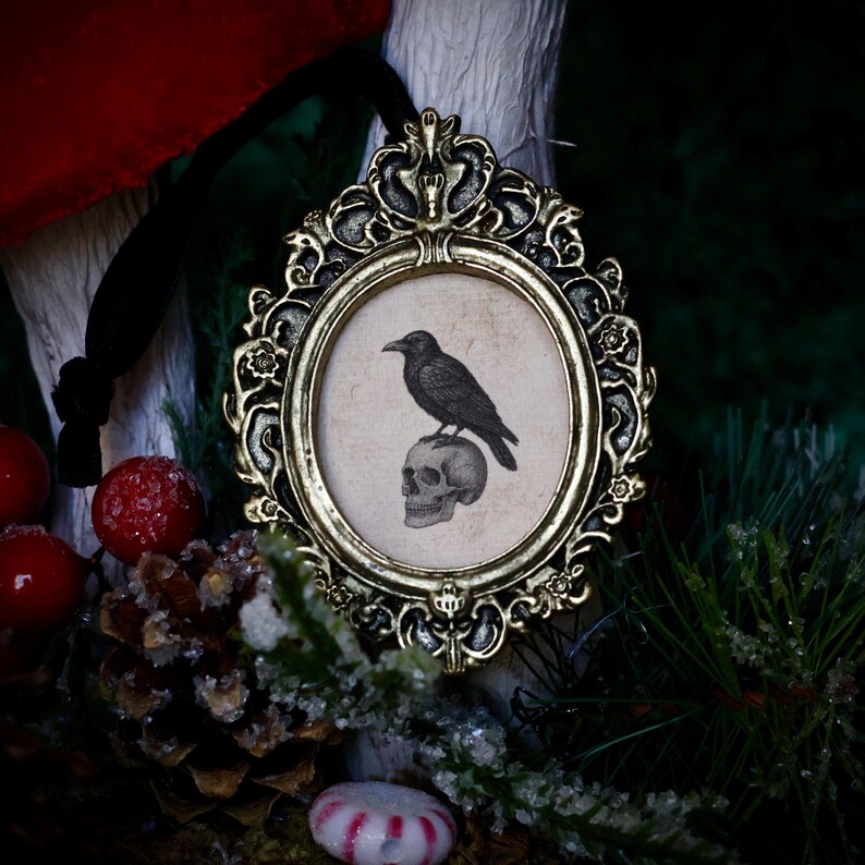 May include: Ornate gold-colored framed ornament featuring a black raven perched atop a skull. The oval image has a vintage aesthetic. A black ribbon is attached for hanging. The background includes red berries, pine needles, and a pine cone.