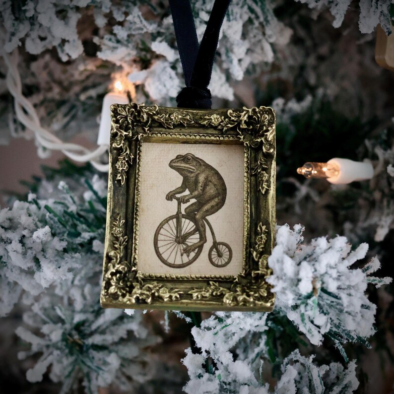 May include: A vintage-style Christmas ornament featuring a framed illustration of a frog riding a penny-farthing bicycle. The frame is ornate with a gold finish, and the ornament hangs from a black ribbon. The background is a snow-covered Christmas tree.