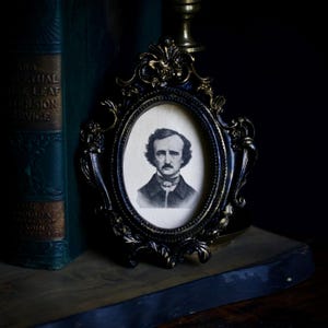 May include: An oval-shaped picture frame with an ornate, dark black and gold finish containing a black and white portrait of Edgar Allan Poe. The frame features intricate scrollwork and floral detailing along the edges, surrounding the central image.