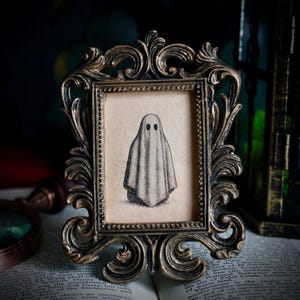 Peut inclure: Une impression encadr&eacute;e de style vintage repr&eacute;sentant un fant&ocirc;me simple. L'&oelig;uvre d'art est en noir et blanc sur un fond beige, dans un cadre dor&eacute; antique orn&eacute; de volutes d&eacute;taill&eacute;es.
