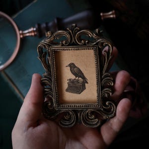 Puede incluir: Marco de fotos ornamentado de estilo antiguo, con un acabado detallado en bronce oscuro. El marco contiene una pequeña impresión de un cuervo posado sobre una máquina de escribir vintage. La obra de arte está sobre un fondo beige.