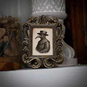 May include: Ornate, antique gold-colored picture frame with a black and white illustration of a plague doctor. The frame has a detailed, scrolling design. The illustration depicts a figure in a long coat and hat with a bird-like mask.