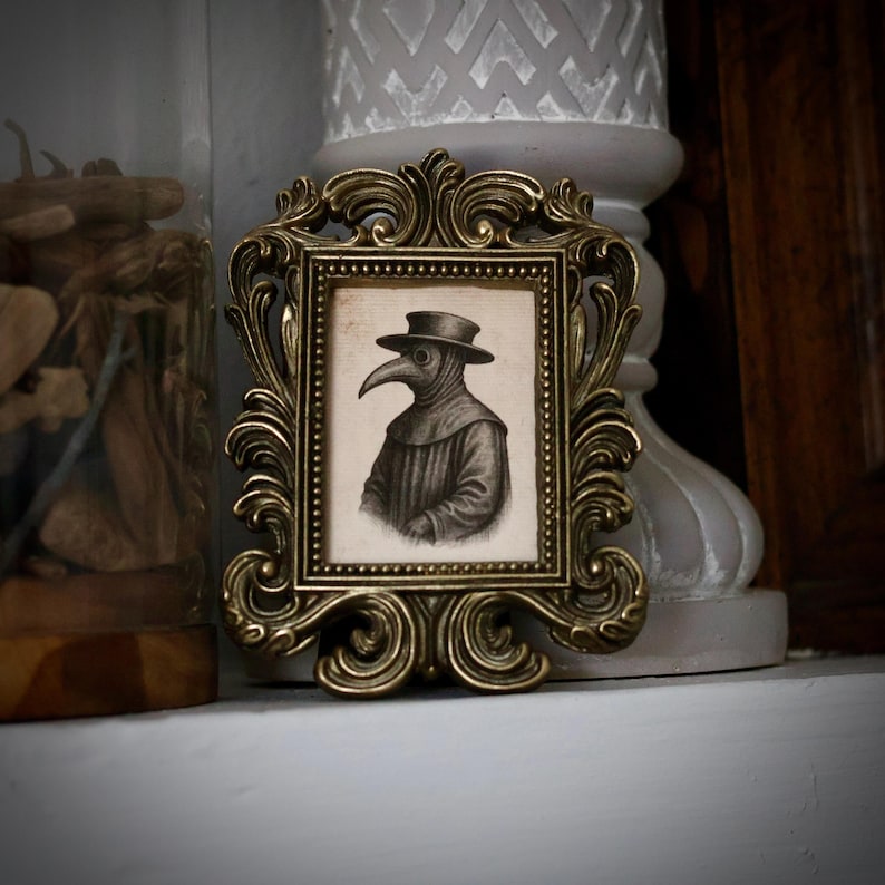 May include: Ornate, antique gold-colored picture frame with a black and white illustration of a plague doctor. The frame has a detailed, scrolling design. The illustration depicts a figure in a long coat and hat with a bird-like mask.