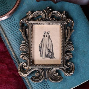 Peut inclure: Cadre photo de style antique orn&eacute; d'un motif tourbillonnant d&eacute;taill&eacute; dans une finition m&eacute;tallique. Le cadre contient une illustration de style vintage d'un fant&ocirc;me portant un n&oelig;ud. L'&oelig;uvre est en noir et blanc.
