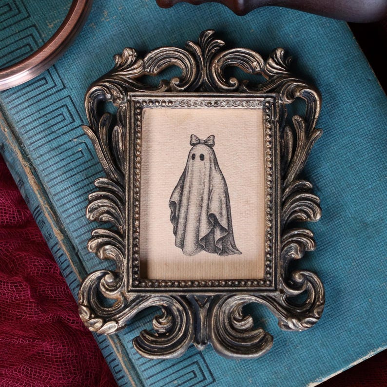 Peut inclure: Cadre photo de style antique orn&eacute; d'un motif tourbillonnant d&eacute;taill&eacute; dans une finition m&eacute;tallique. Le cadre contient une illustration de style vintage d'un fant&ocirc;me portant un n&oelig;ud. L'&oelig;uvre est en noir et blanc.
