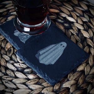 May include: Two black slate coasters with a ghost design. A glass of red liquid sits on top of one coaster. The coasters are rectangular and have a natural, uneven edge. The ghost design is a simple, cartoon-like illustration.