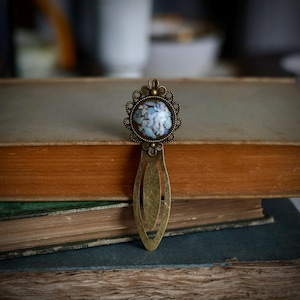 Könnte beinhalten: Ein Lesezeichen aus Messing im Vintage-Stil mit ovalem Design. Der obere Teil zeigt eine runde Verzierung mit einem blau-weißen Blumenmuster, umrahmt von dekorativem Filigran. Das Lesezeichen liegt auf einem Stapel alter Bücher.