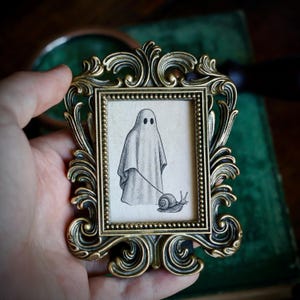 May include: A small, ornate, antique gold-colored picture frame holds a black and white drawing of a ghost walking a snail on a leash. The frame has an elaborate, swirling design.