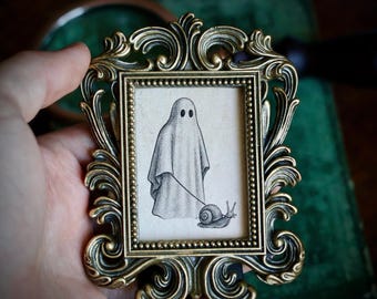 Può includere: Una piccola cornice dorata ornata contiene un disegno in bianco e nero di un fantasma che porta a spasso una lumaca al guinzaglio. La cornice presenta un elaborato design a volute. L'opera d'arte è su uno sfondo color crema.